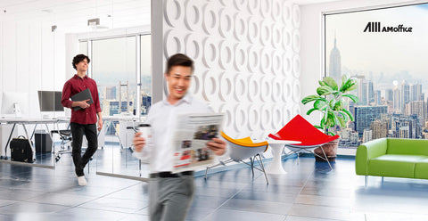 A white themed office with lego style wall in between a working area and a lounge area. Two office workers are walking around the office as they work